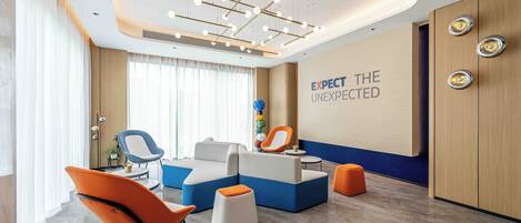 Un área de espera moderna con un sofá modular, sillas anaranjadas y una pared con el texto 'EXPECT THE UNEXPECTED'.