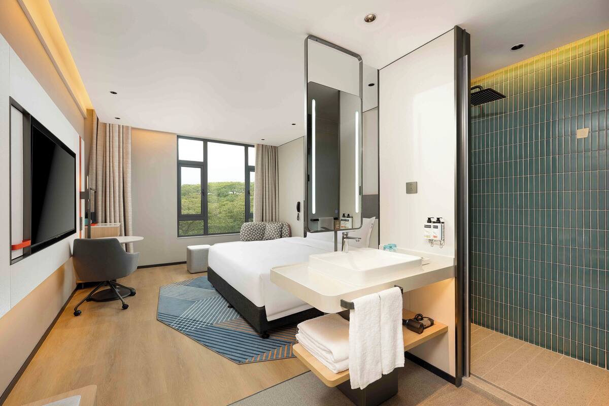 Habitación de hotel moderna con una cama grande, un escritorio, un televisor de pantalla plana, un ba ño con un espejo amplio y vistas a zonas verdes.