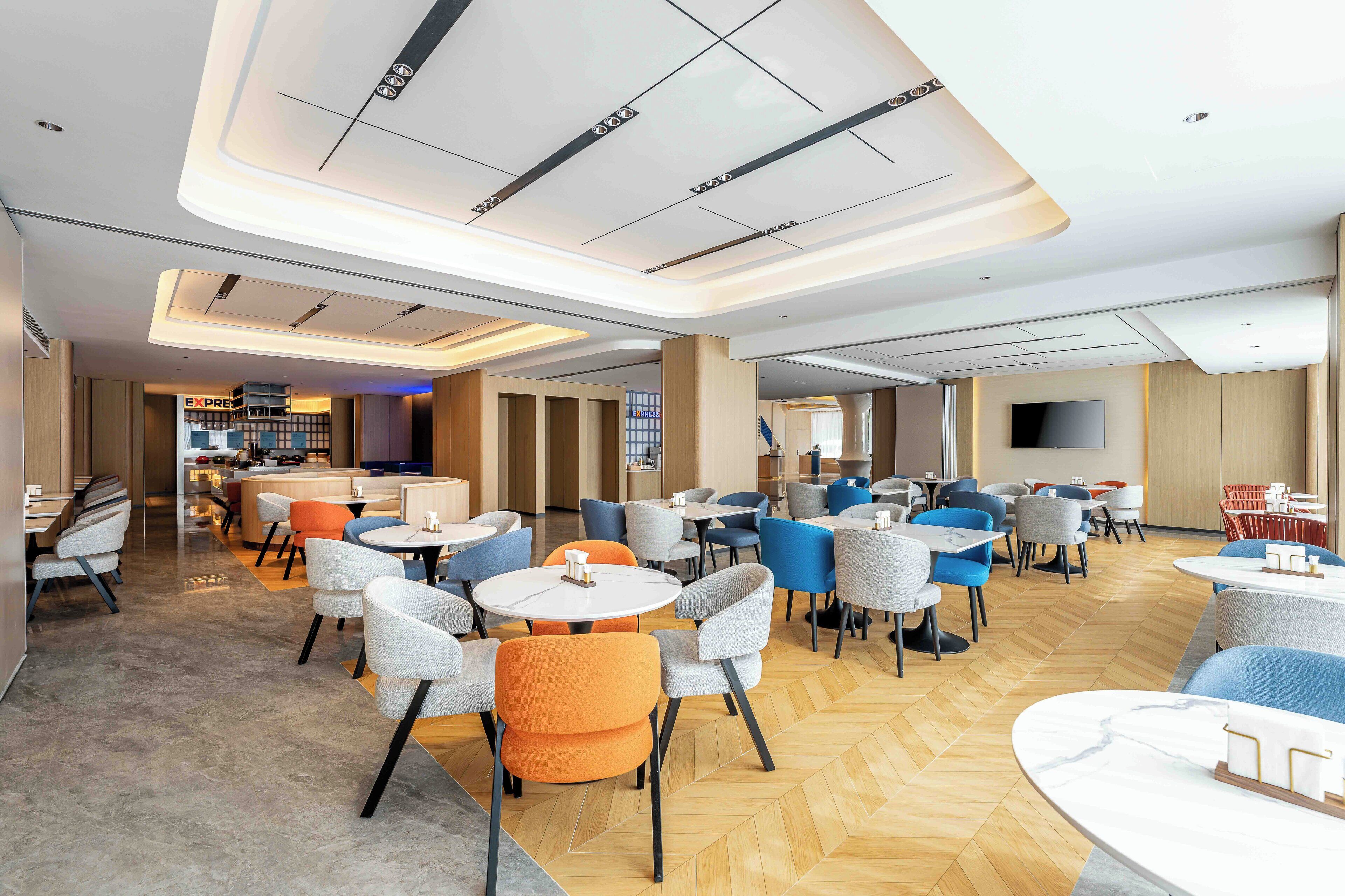 En moderne hotellobby med forskellige siddepladser, herunder runde borde og stole, samt et centralt område med disk og fjernsynsskærm.