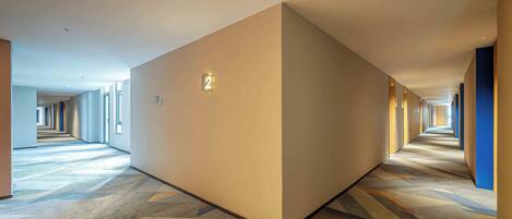 Un largo y estrecho pasillo de hotel con suelo de alfombra, un cartel con número en la pared y una puerta a la derecha.