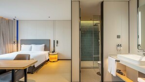 Una habitación de hotel moderna con cama, mesita de noche, lámpara y baño con lavamanos y ducha.