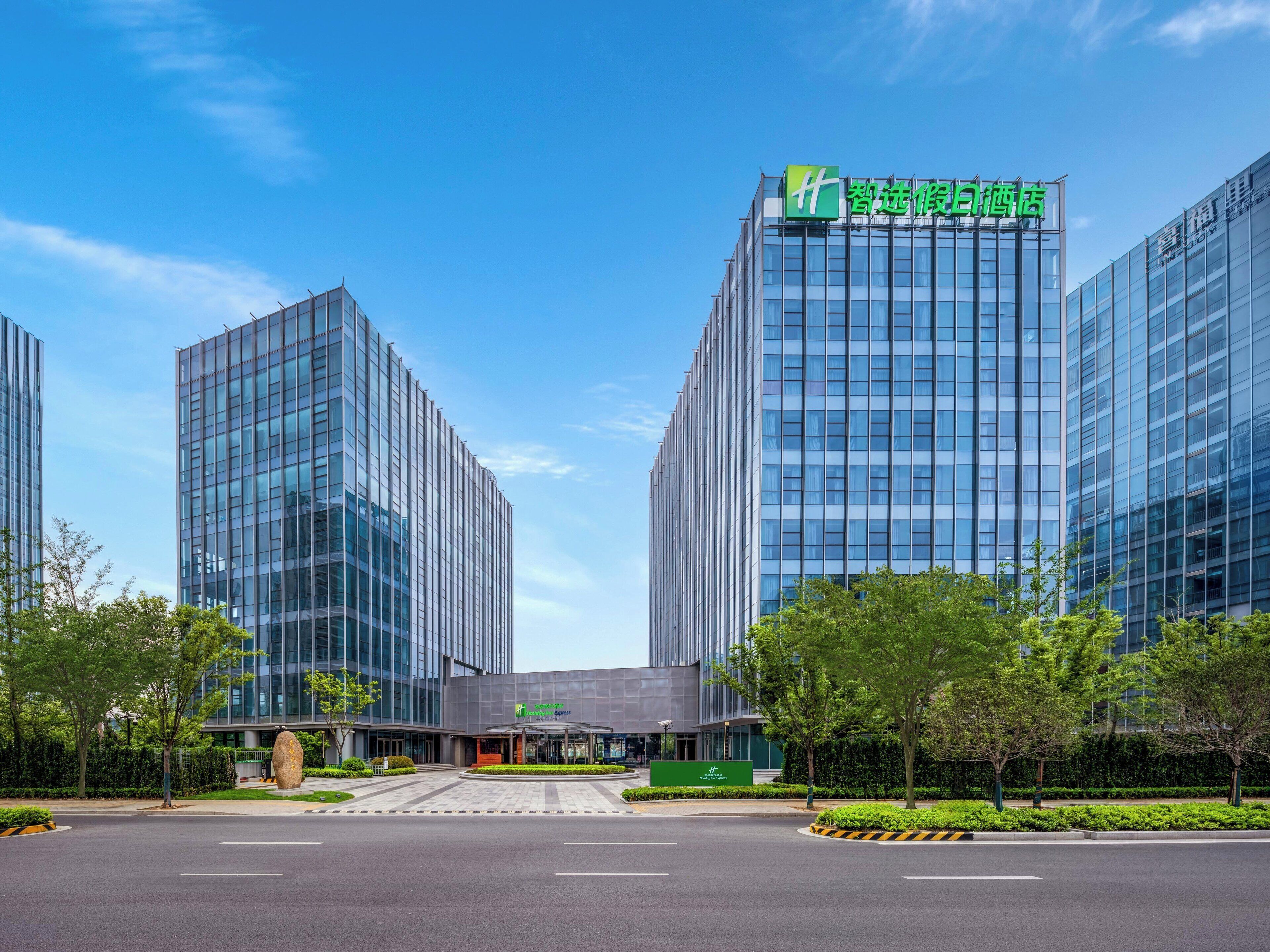 A modern hotel with a green sign, large glass windows, and a well-maintained garden area.
