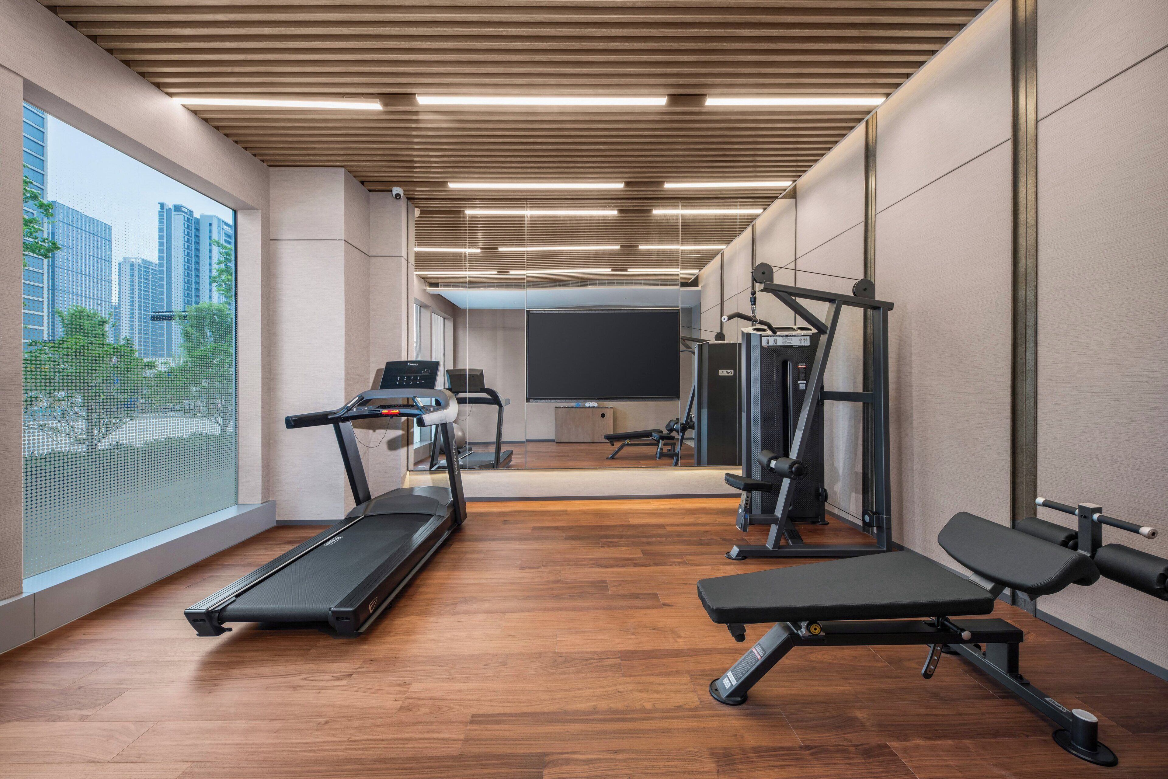 A modern gym with a treadmill, bench press, and weight machines, large windows with a city view, and a wooden ceiling.