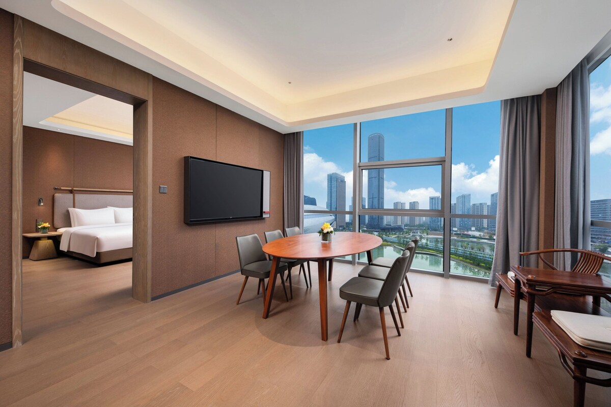 A modern hotel room with a large window, a flat-screen TV, a round dining table, and a bed.