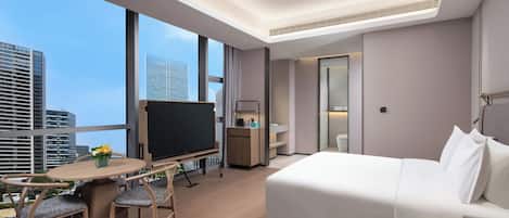 A modern hotel room with a large bed, a desk with a flat-screen TV, a dining area with chairs, and a view of the city skyline.