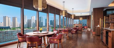 A modern dining area with wooden tables and chairs, large windows with a city view, and hanging pendant lights.