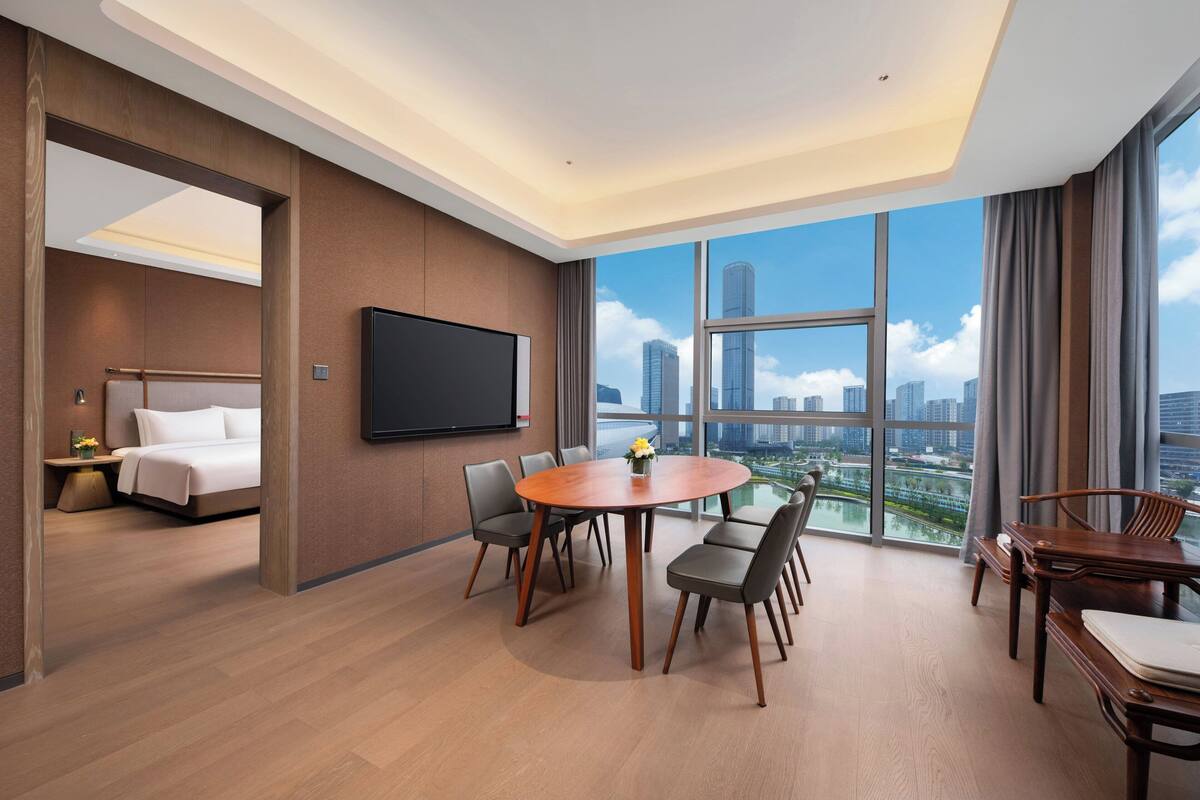 Habitación de hotel moderna con un ventanal grande, una televisión de pantalla plana, una mesa de comedor redonda y una cama.