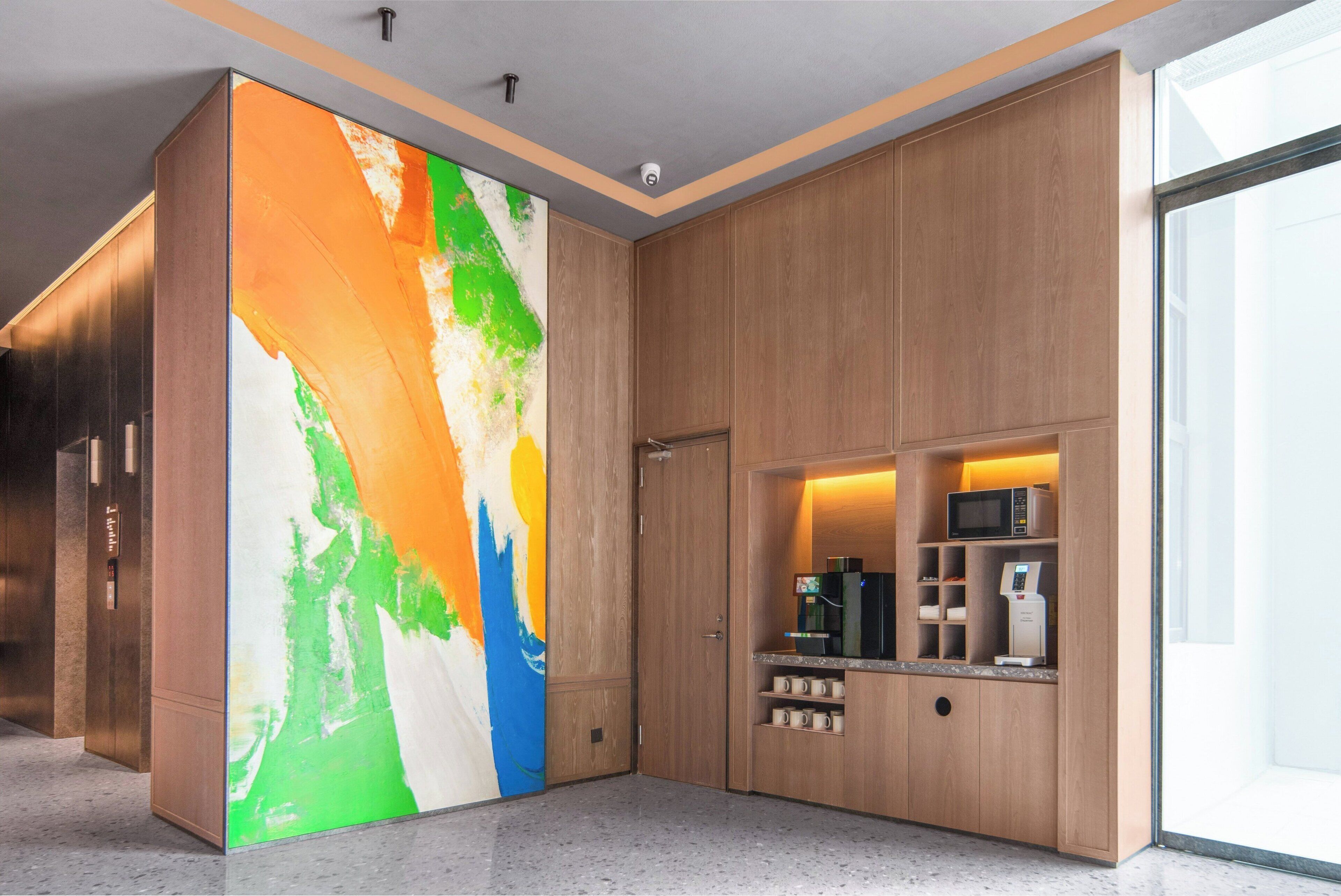 A modern hotel lobby with a large abstract painting, wooden paneling, a coffee station, and a glass door leading to the outside.