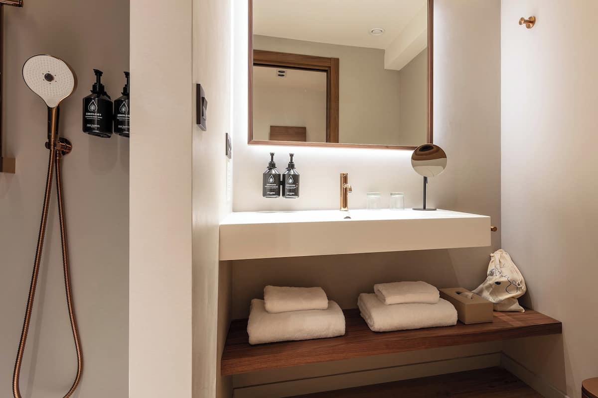 Un baño con un lavamanos blanco, un espejo y dos botellas de gel de ducha Zenology. Hay toallas en un estante y un cabezal de ducha con una manguera de mano.