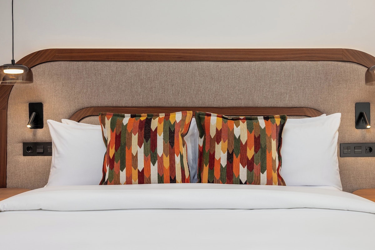 Una cama con ropa de cama blanca, un cabecero estampado y llamativo, y dos lámparas fijadas a la pared.