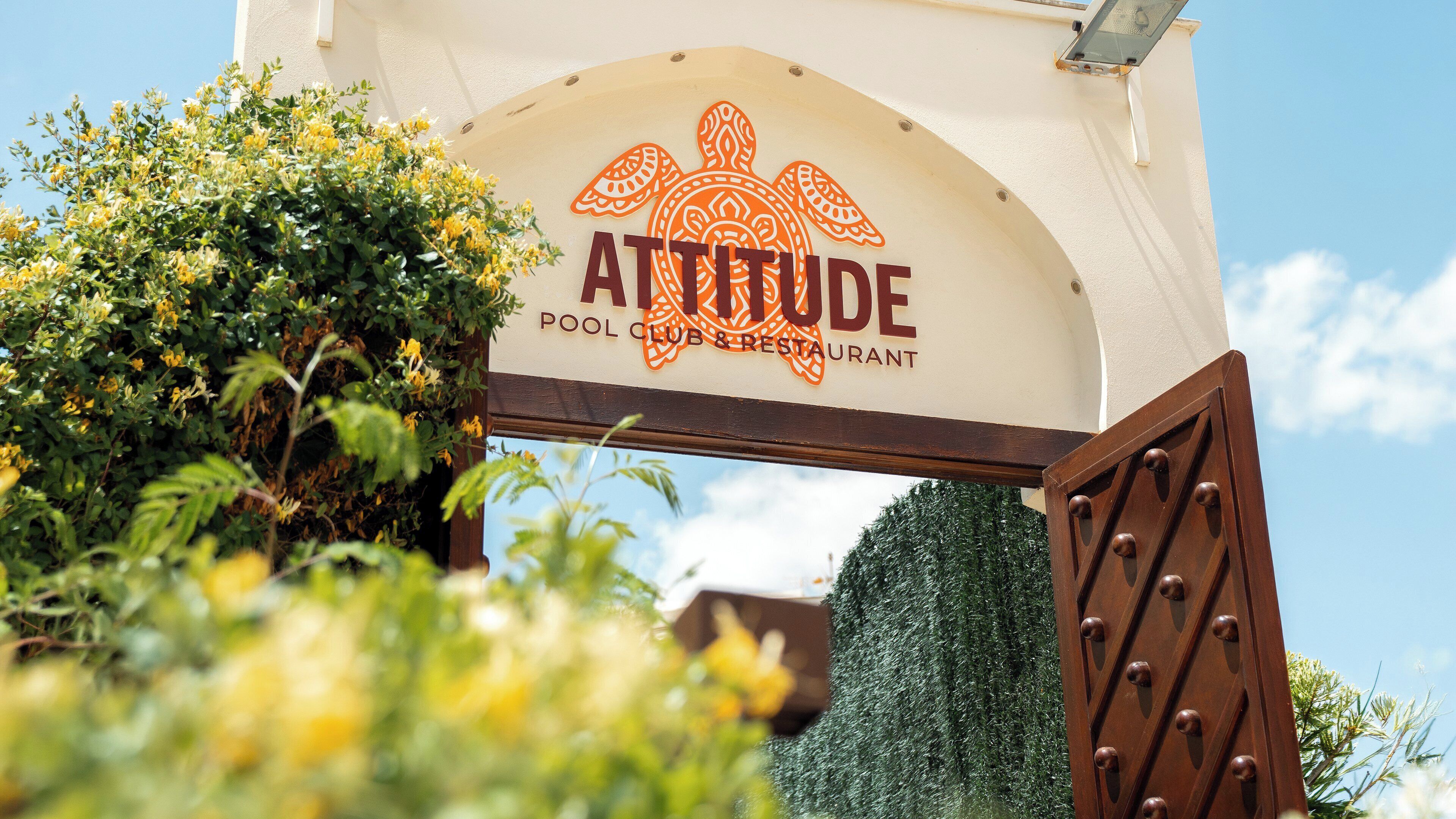 Schild für 'ATTITUDE POOL CLUB & RESTAURANT' mit Schildkröten-Logo, umgeben von Grünpflanzen.