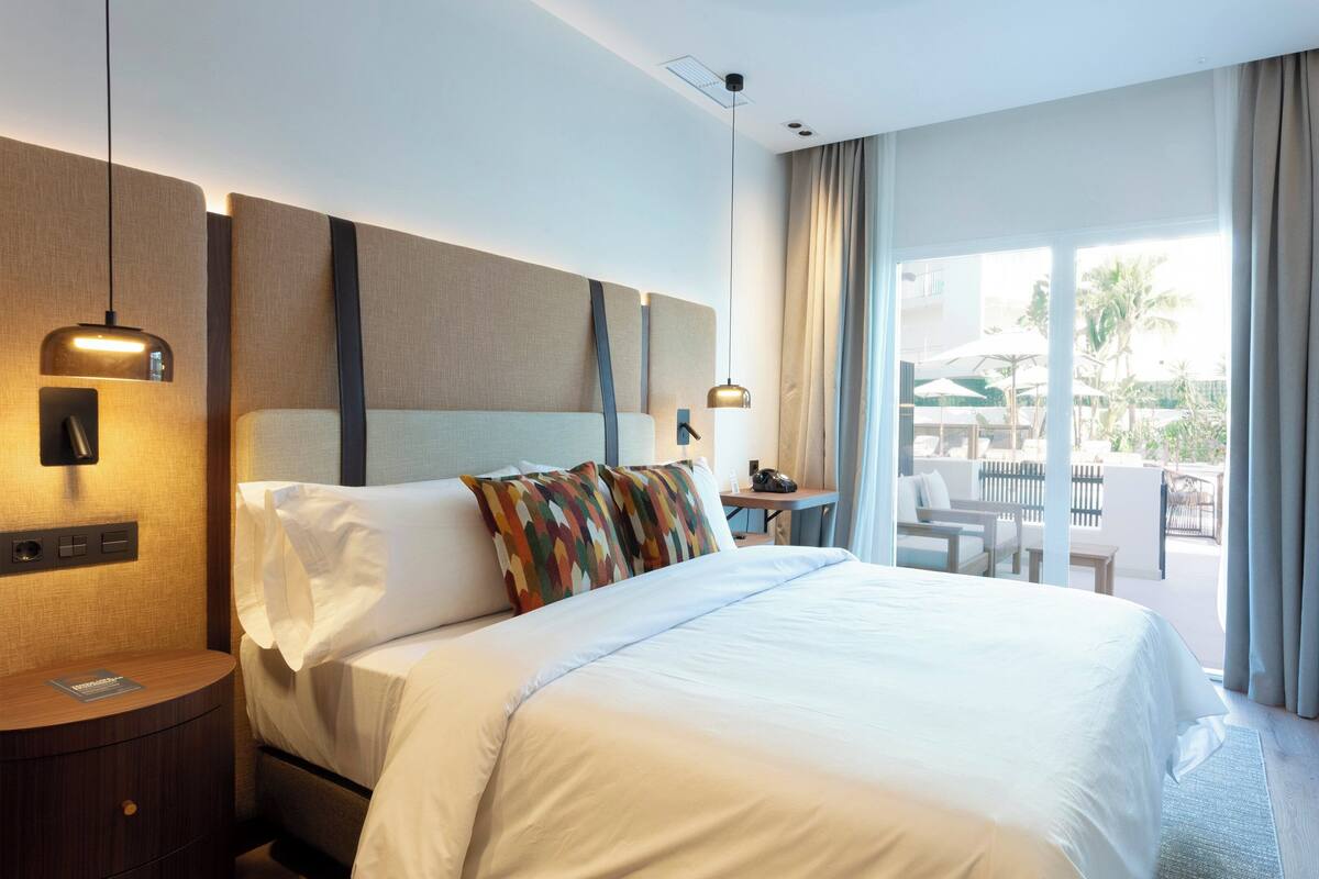 Et hotelværelse med en stor seng, et natbord, et skrivebord med stol, og udsigt til det åbne areal.
