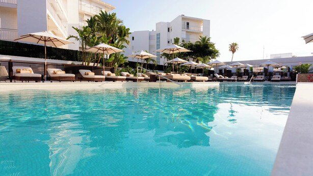 Una piscina con sillas de descanso y sombrillas, rodeada de edificios y palmeras.