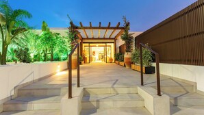 Una entrada exterior bien iluminada con una pérgola de madera, escalones que conducen a un edificio y diversas plantas en macetas.