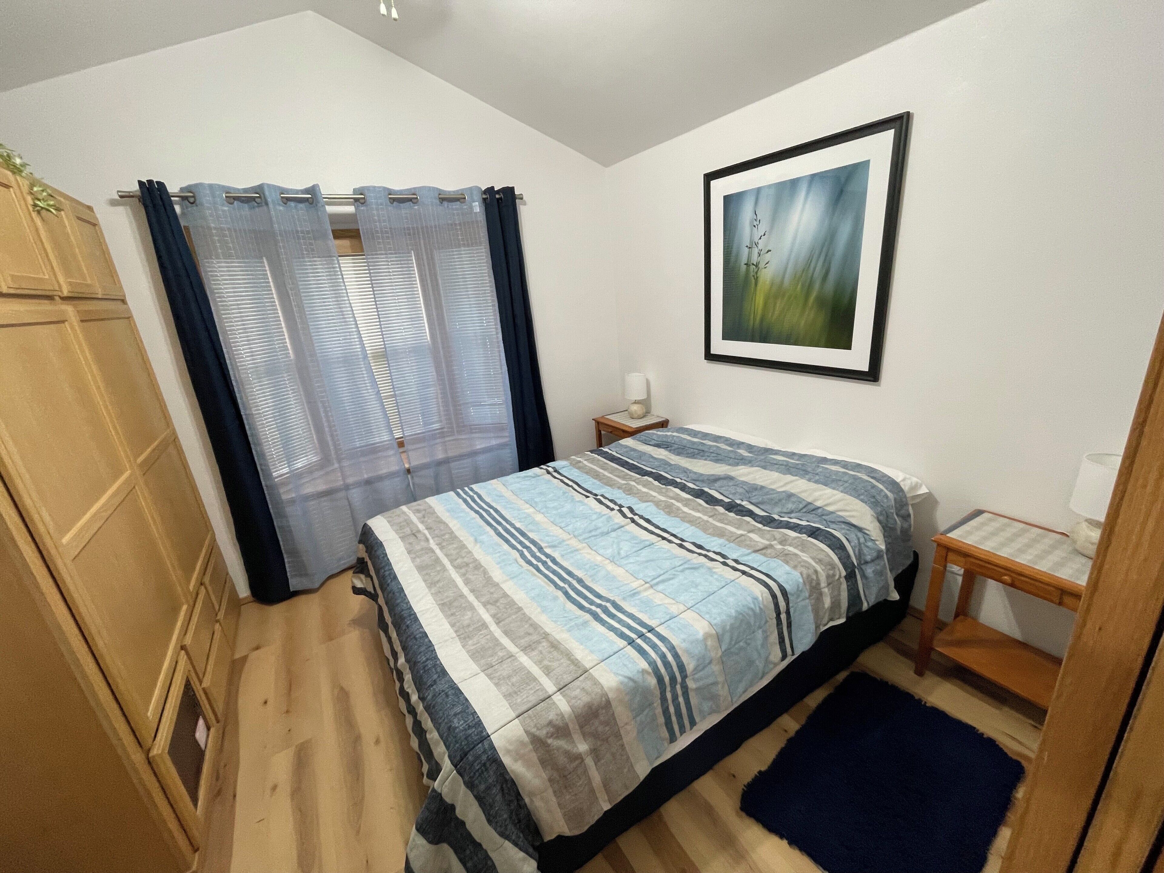Une chambre à coucher avec un lit, une table de chevet, un tableau encadré au mur et une fenêtre avec des rideaux.