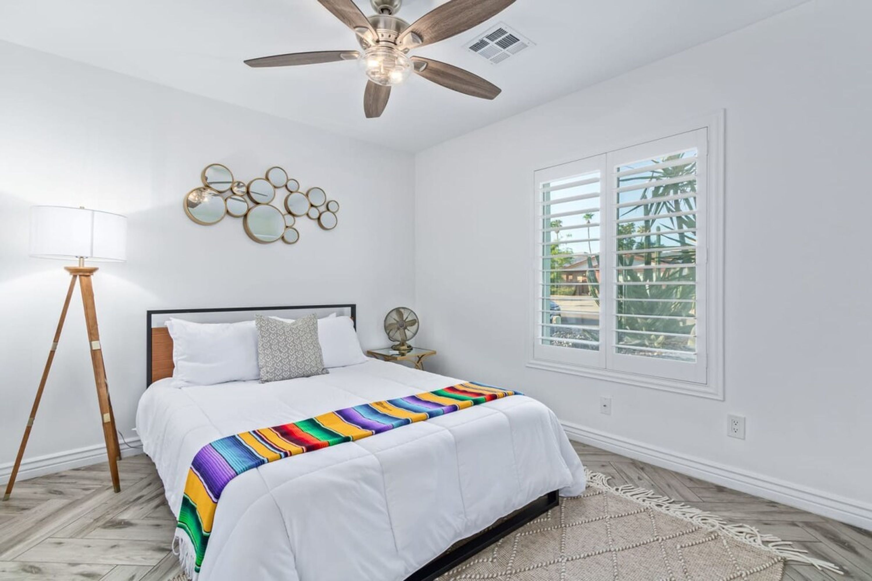 A bedroom with a bed, a ceiling fan, a bedside table with a lamp, a window with blinds, and a wall decoration.