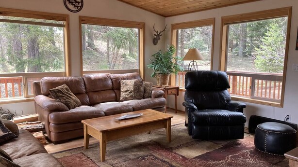 Un salon chaleureux avec un canapé en cuir marron, un fauteuil en cuir noir et une table basse en bois. Une grande fenêtre offre une vue sur les arbres, un mur est décoré d’une tête de cerf et un panier contient des plantes.