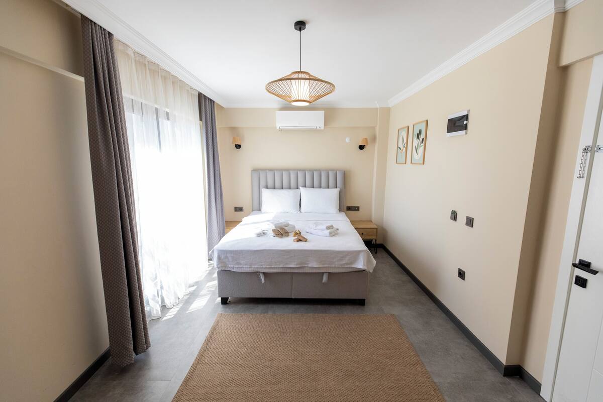 Una habitación de hotel con una cama, mesitas de noche, una lámpara colgante y un ventanal con cortinas.