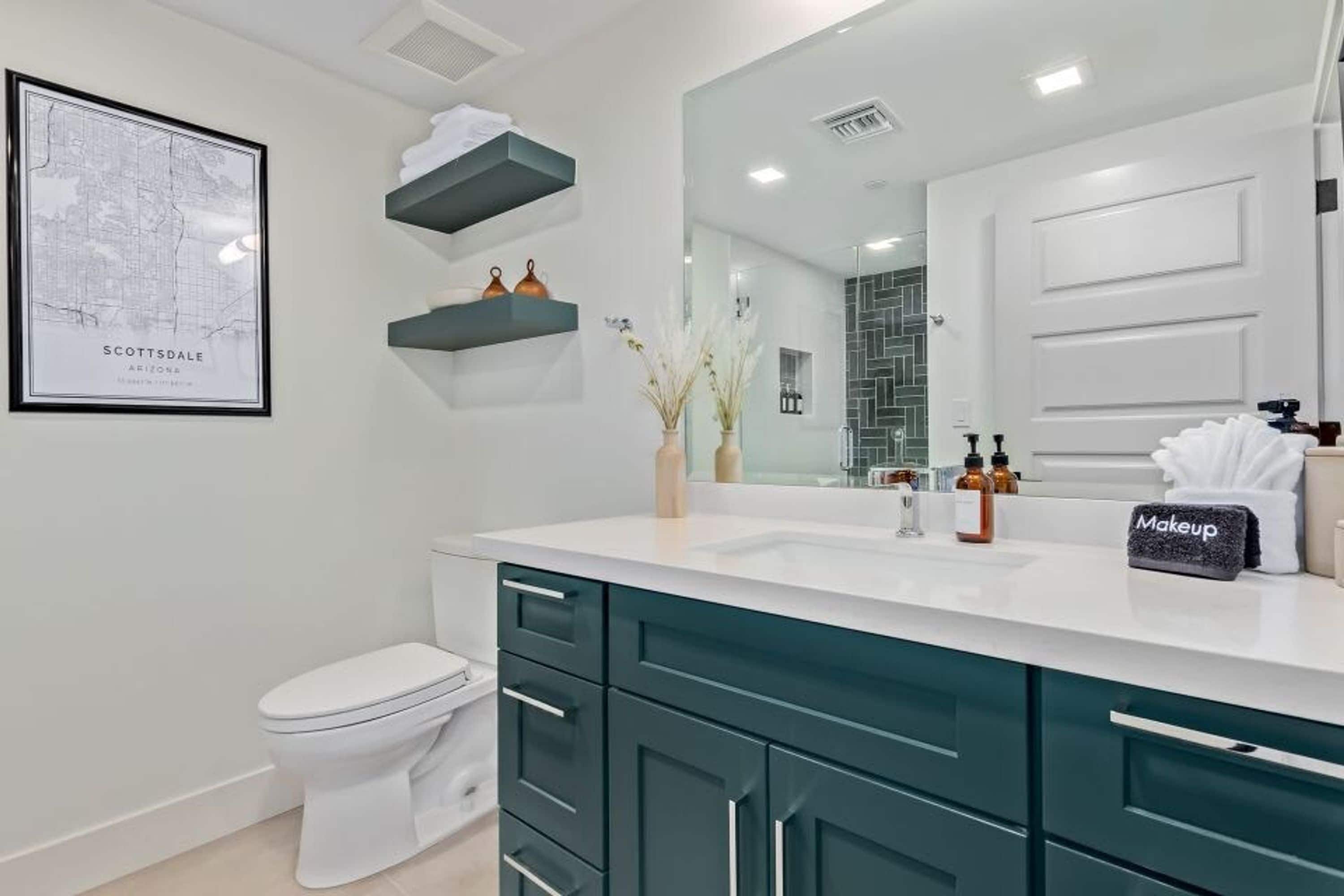 A modern bathroom with a white toilet, teal cabinets, a white sink, and a large mirror. There are decorative items on the counter and a framed map on the wall.