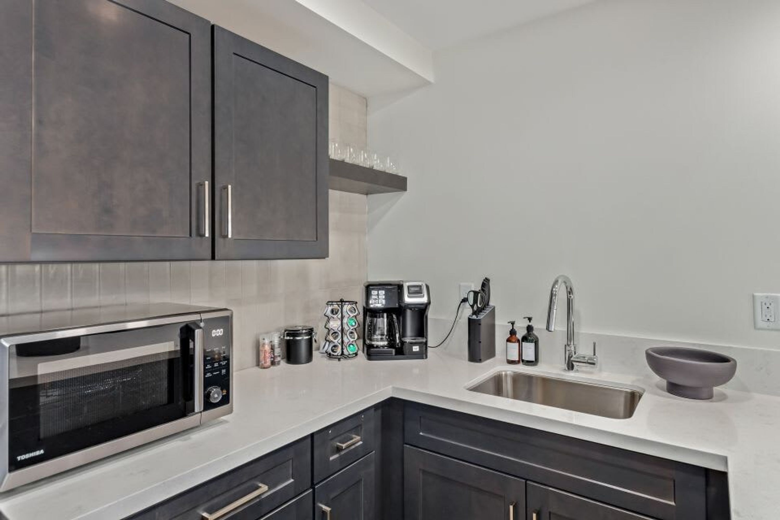 A modern kitchen with dark cabinets, a stainless steel sink, a microwave, a coffee maker, and a bowl on the countertop.