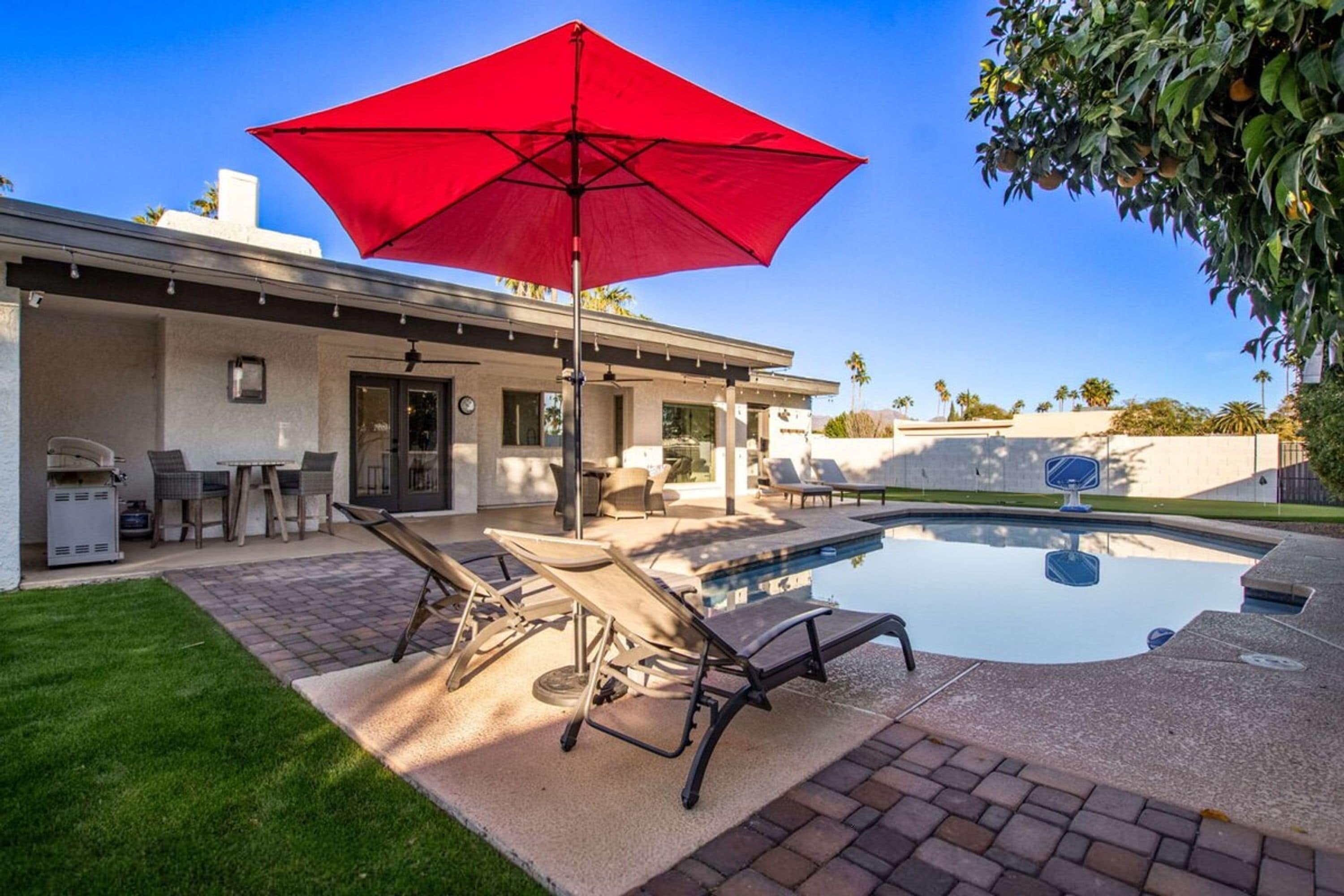 Un jardin avec une piscine, des chaises longues, un parasol rouge et un barbecue.