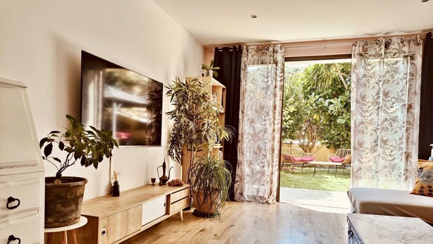 Un salón moderno con suelo de madera, un televisor de pantalla plana grande montado en la pared, una mesa consola de madera con plantas y un ventanal con cortinas.
