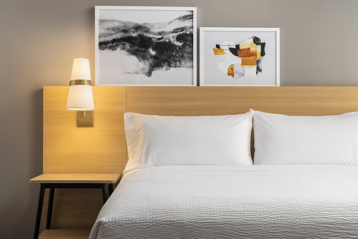 Ein Hotelzimmer mit hölzernem Kopfteil, weißer Bettwäsche, einem Nachttisch mit Lampe und zwei gerahmten abstrakten Kunstwerken an der Wand.