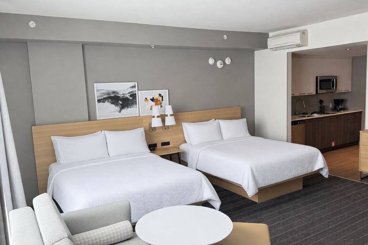 Ein Hotelzimmer mit zwei Betten, einem kleinen Tisch, einer Couch, einer Küchenzeile und Bildern an der Wand.