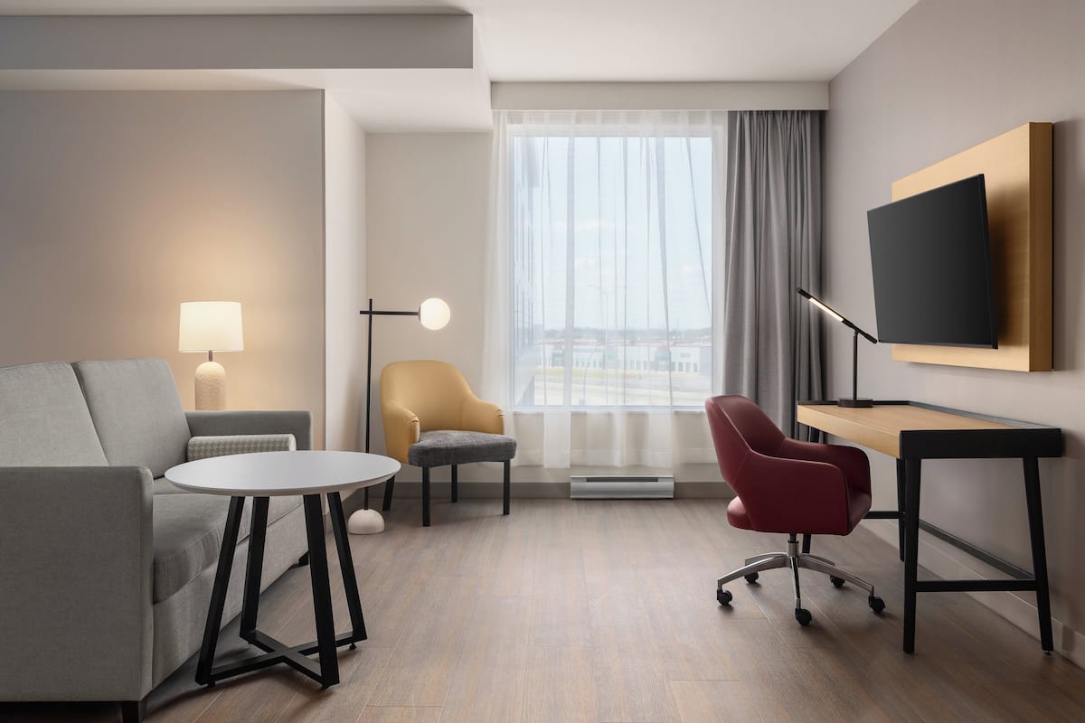 Ein modernes Hotelzimmer mit grauem Sofa, einem kleinen runden Tisch, einem roten Bürostuhl, einem Schreibtisch, einem an der Wand montierten Flachbildfernseher und einem großen Fenster mit Vorhängen.