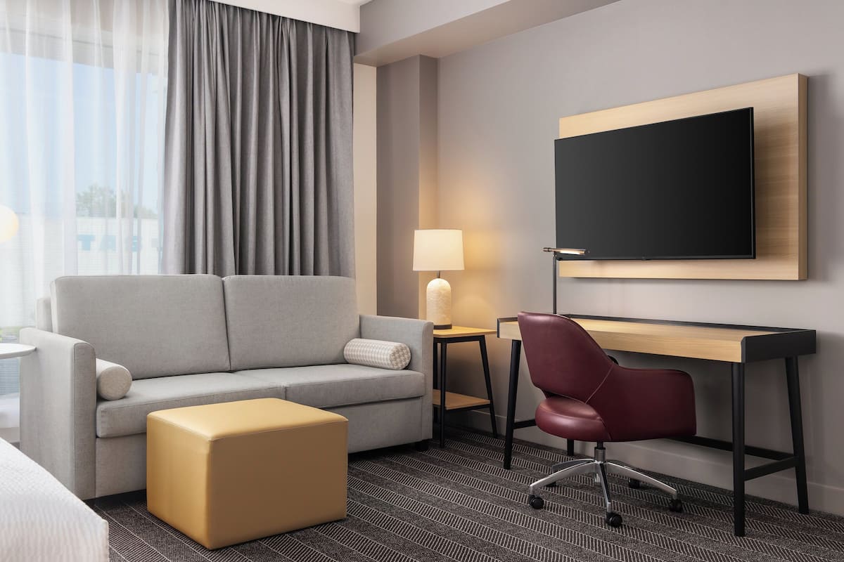 Ein modernes Hotelzimmer mit grauem Sofa, einem Holztisch, einem roten Bürostuhl, einem kleinen Tisch mit Lampe und einem großen Flachbildfernseher an der Wand.