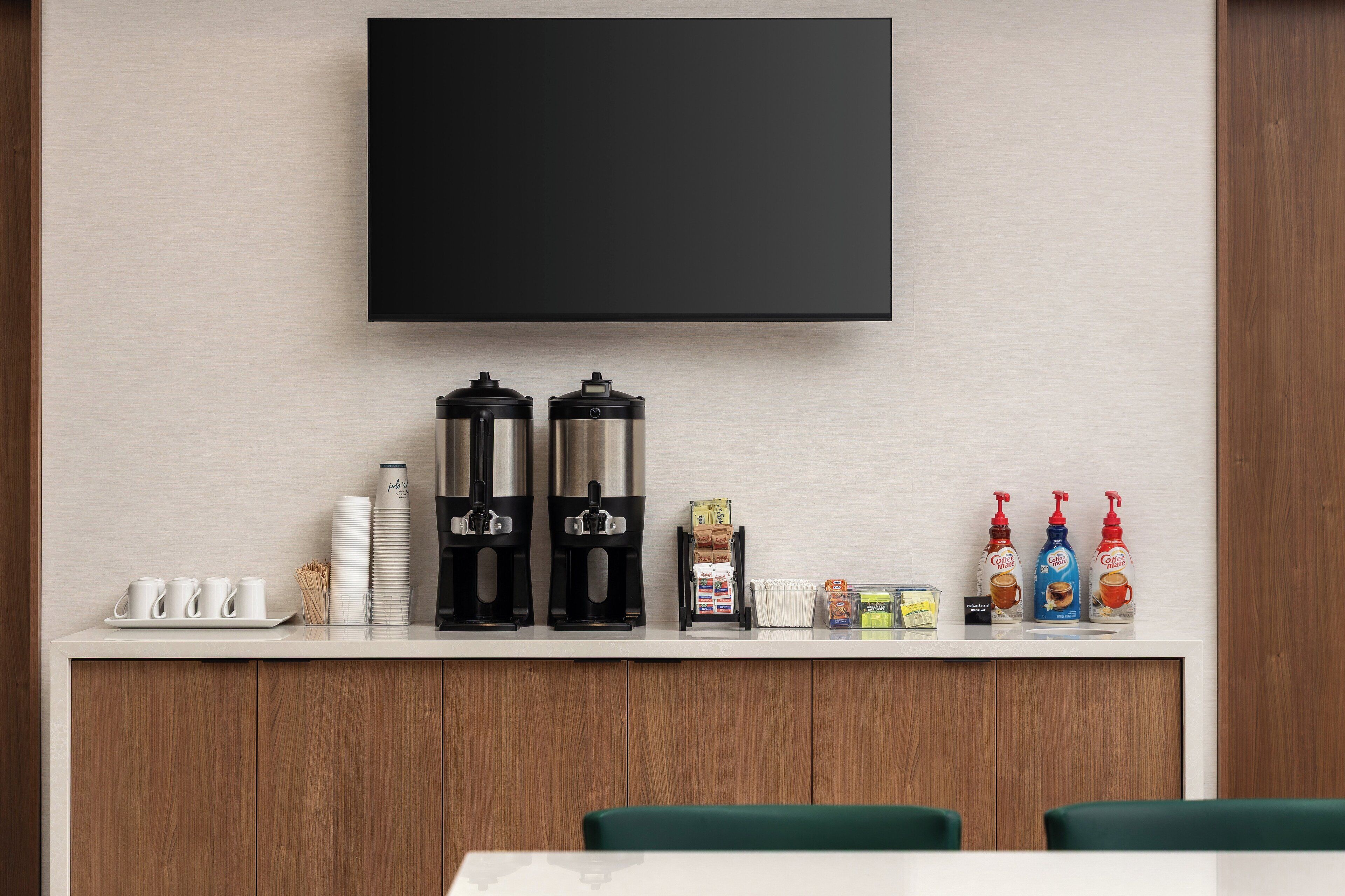 Una habitación de hotel moderna con un televisor de pantalla plana montado en la pared, una estación de café con dos cafeteras, una pila de tazas blancas y una pequeña selección de productos para el desayuno.