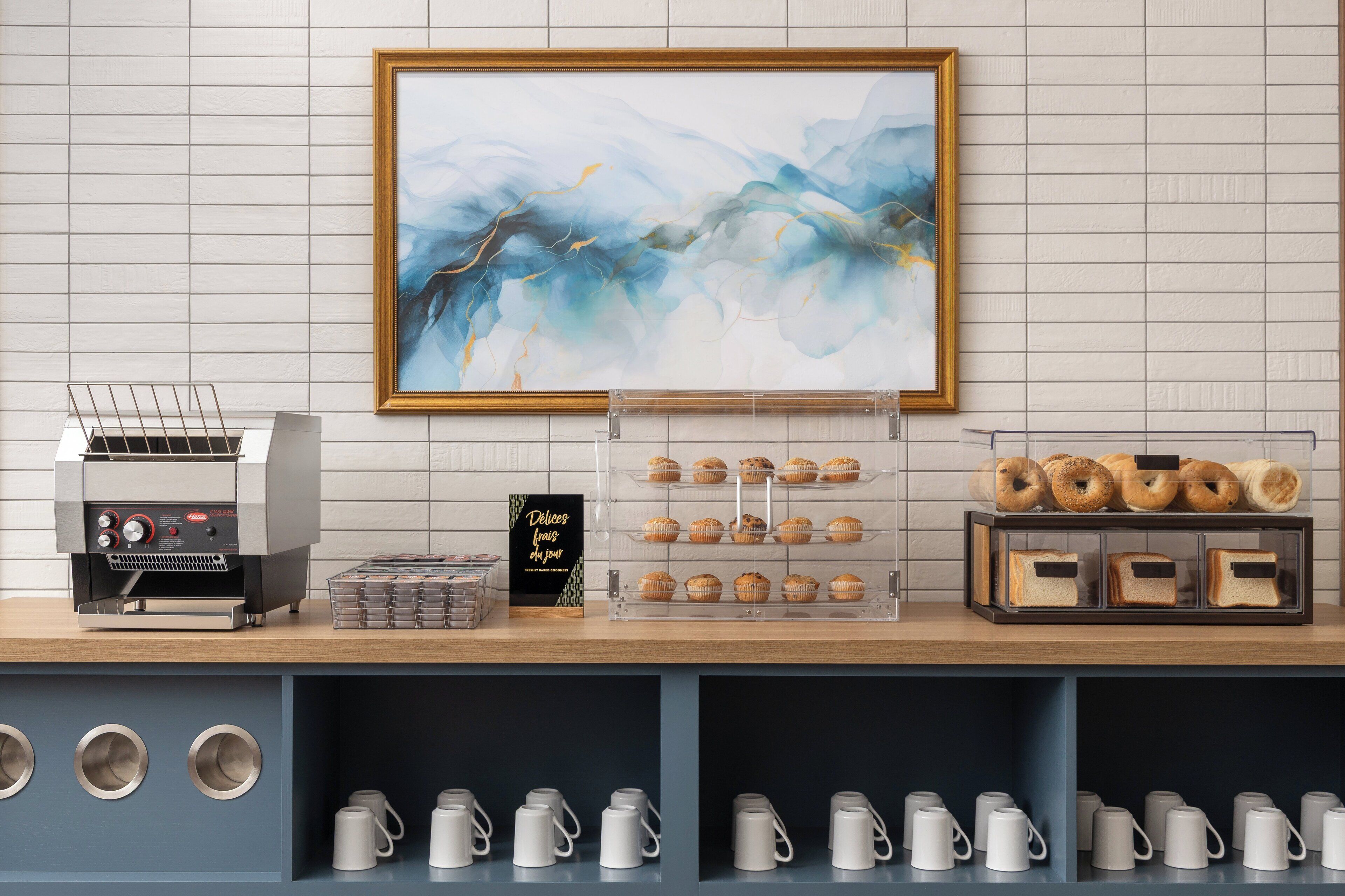 A bakery display with pastries, a coffee machine, and a framed abstract painting on the wall.