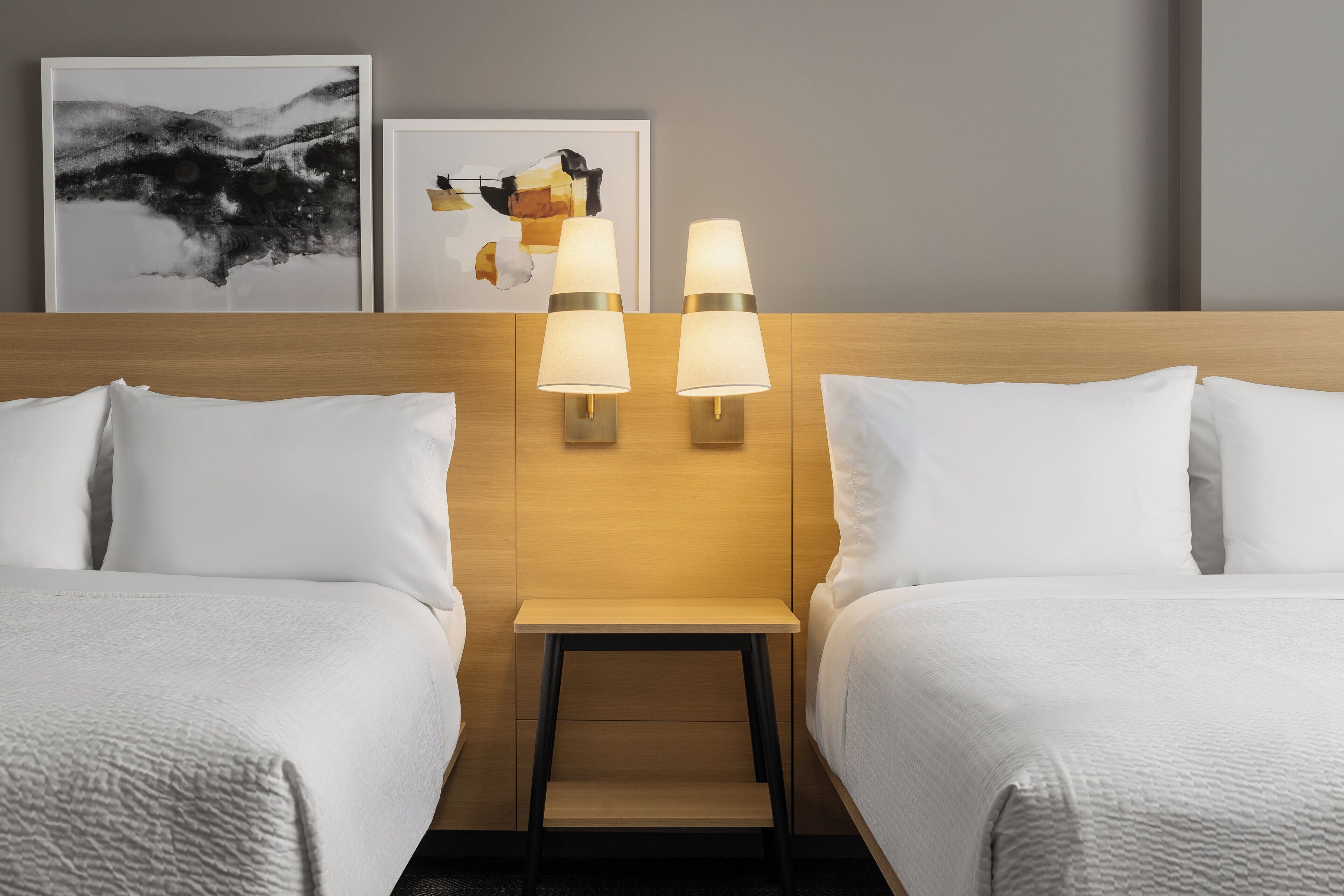 Quarto de hotel com duas camas, uma mesinha de cabeceira e luminárias fixadas na parede.