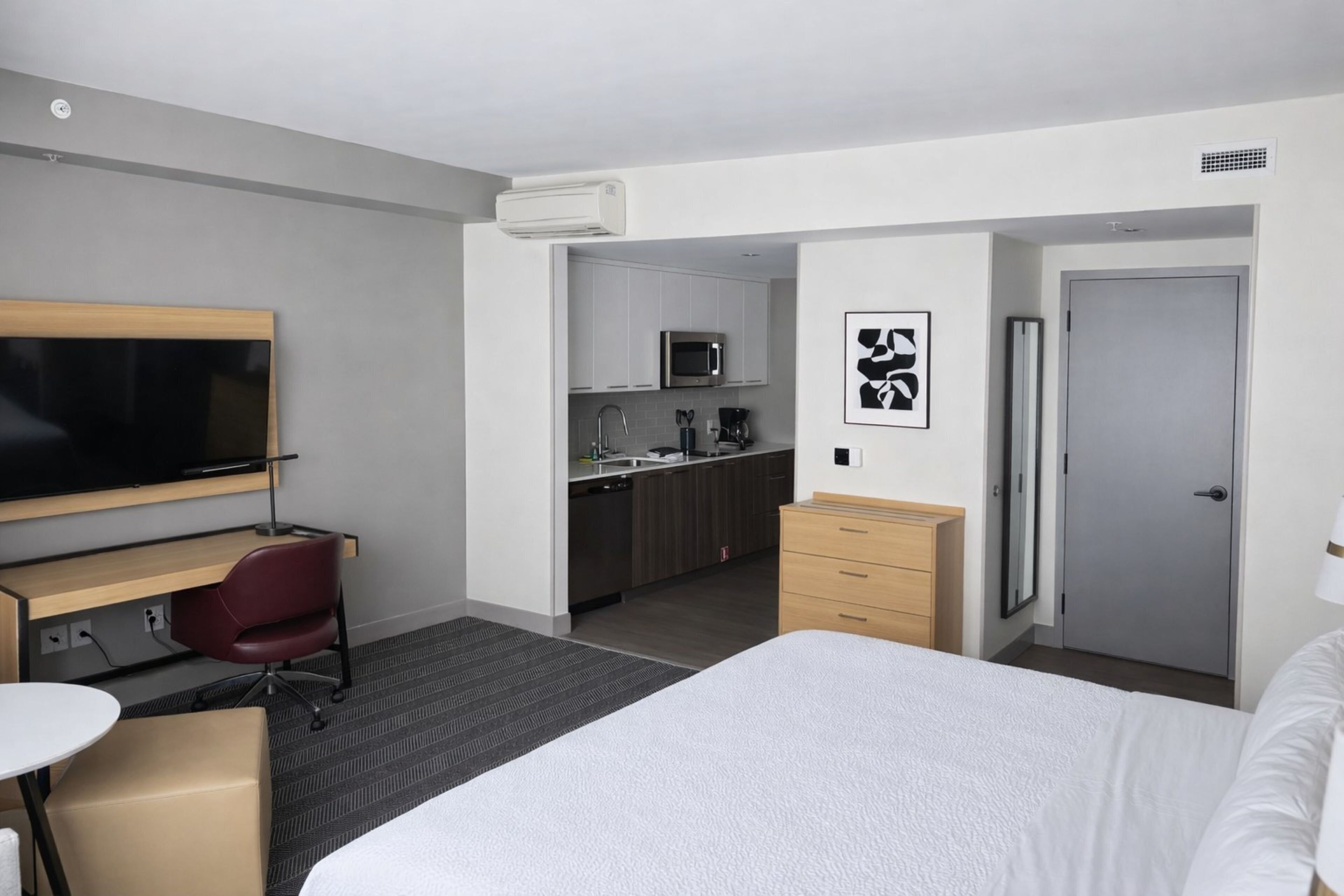A hotel room with a bed, a desk with a chair, a television, a kitchenette, and a door.