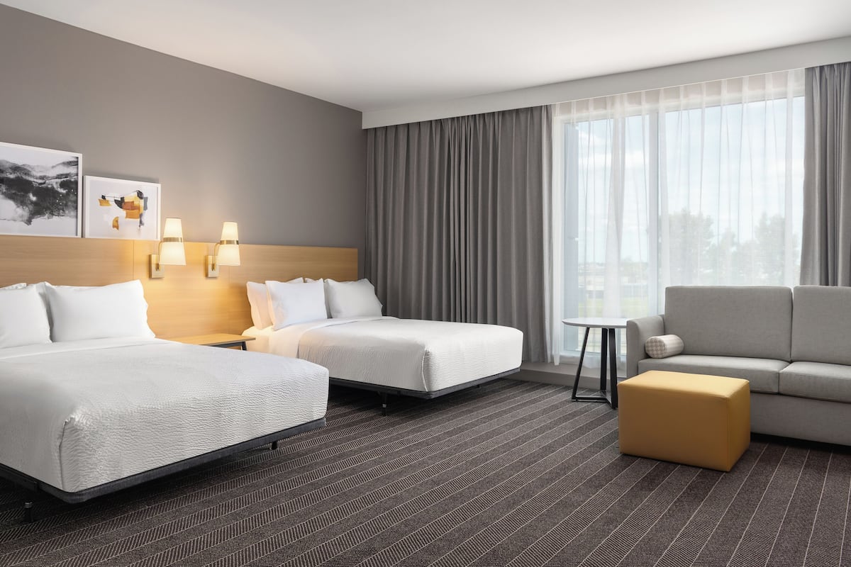 Ein Hotelzimmer mit zwei Betten, einem Schlafsofa, einem kleinen Tisch und einem großen Fenster mit Vorhängen.