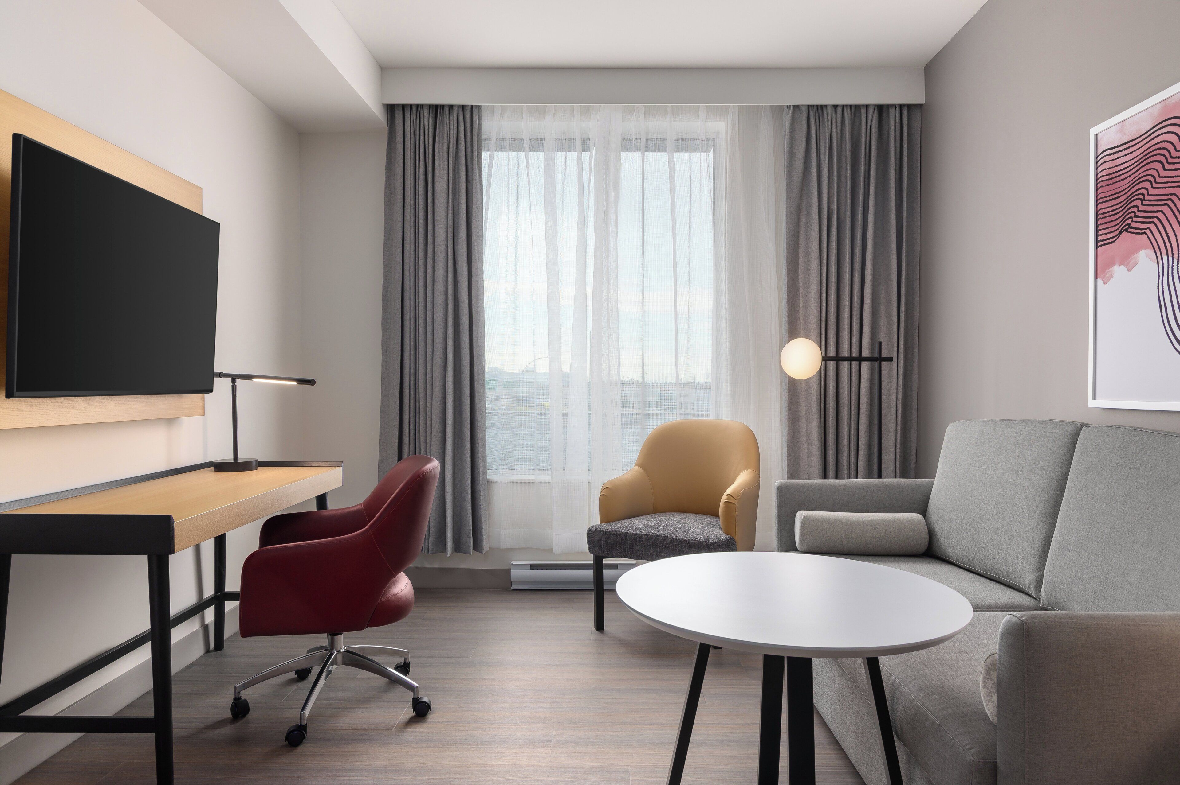 A modern hotel room with a flat-screen TV, a desk, a red office chair, a grey sofa, a small round table, and a large window with curtains.