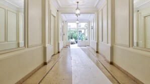 A long, narrow hallway with white walls and a beige floor, leading to an open area with a view of greenery. - A Peaceful Retreat in Le Marais (Paris)