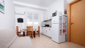 Ein kompakter Raum mit einem Bett, einem Esstisch mit Stühlen, einer Küchenzeile inklusive Kühlschrank, Mikrowelle und Herd sowie einer Holztür.