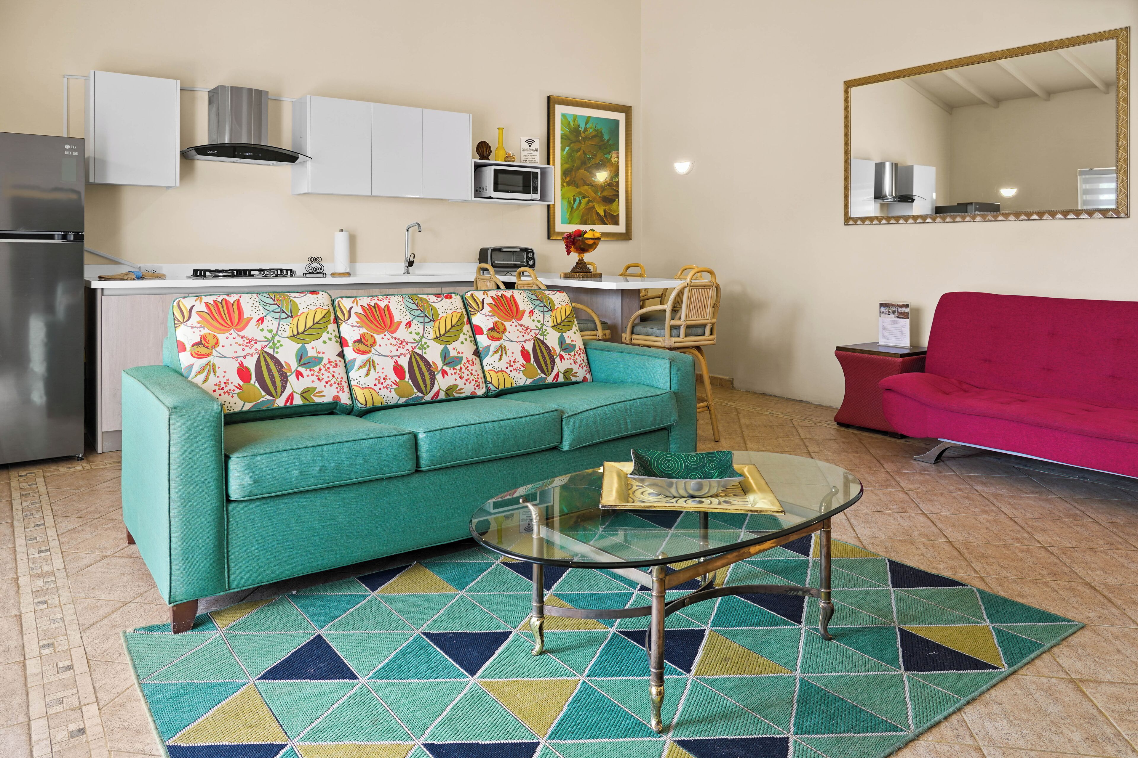 A compact living space with a teal sofa, a glass coffee table, a patterned rug, a kitchen area with white cabinets, a microwave, and a red couch.