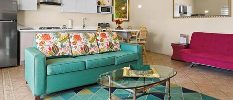 A compact living space with a teal sofa, a glass coffee table, a patterned rug, a kitchen area with white cabinets, a microwave, and a red couch.