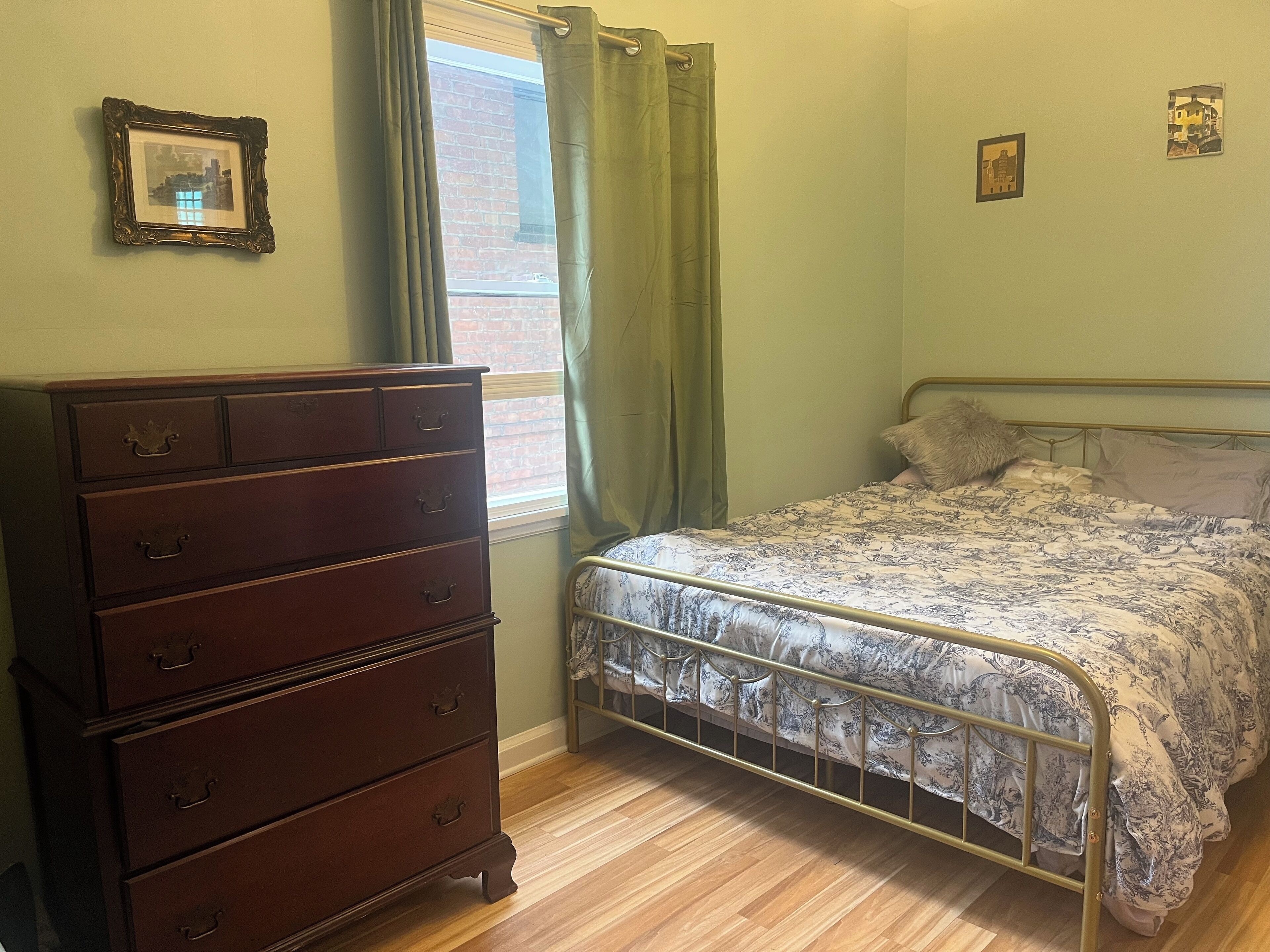 A bedroom with a wooden dresser, a metal bed frame, a patterned bedspread, and a window with curtains.