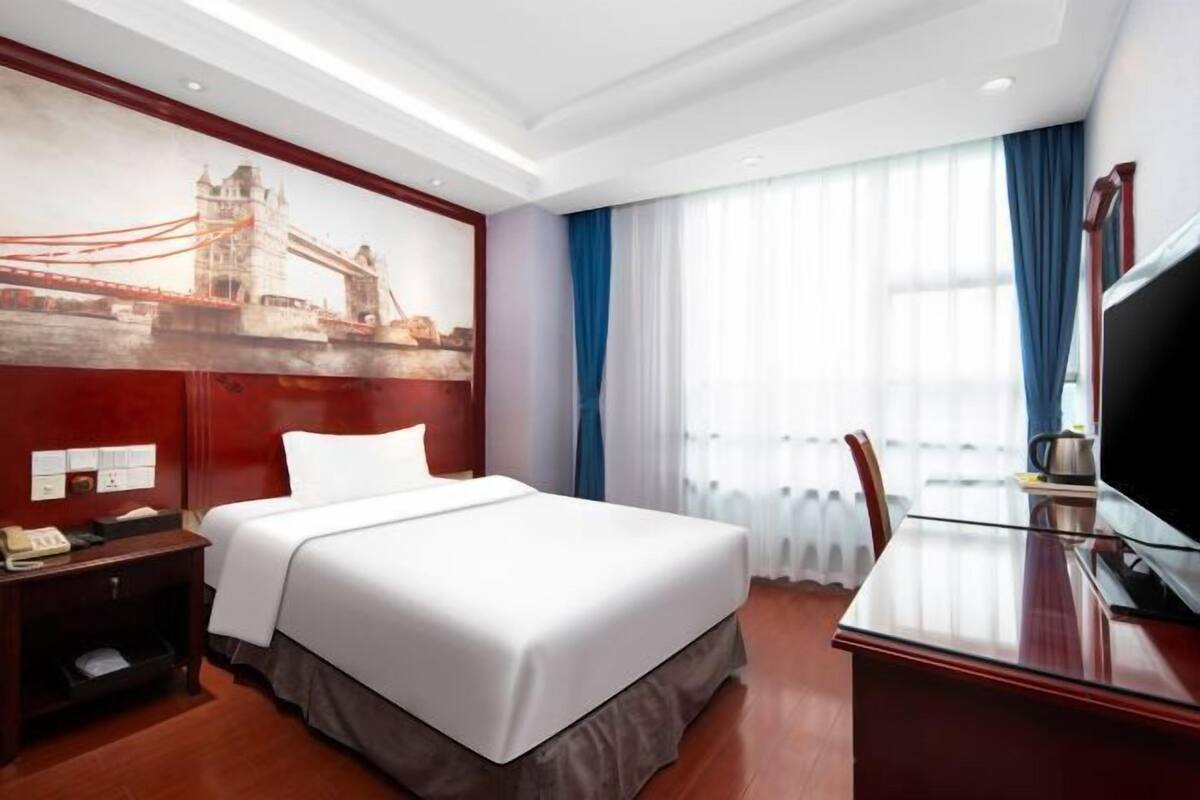 Habitación de hotel con una cama grande, un escritorio con televisor, un cuadro de un puente y una ventana con cortinas.
