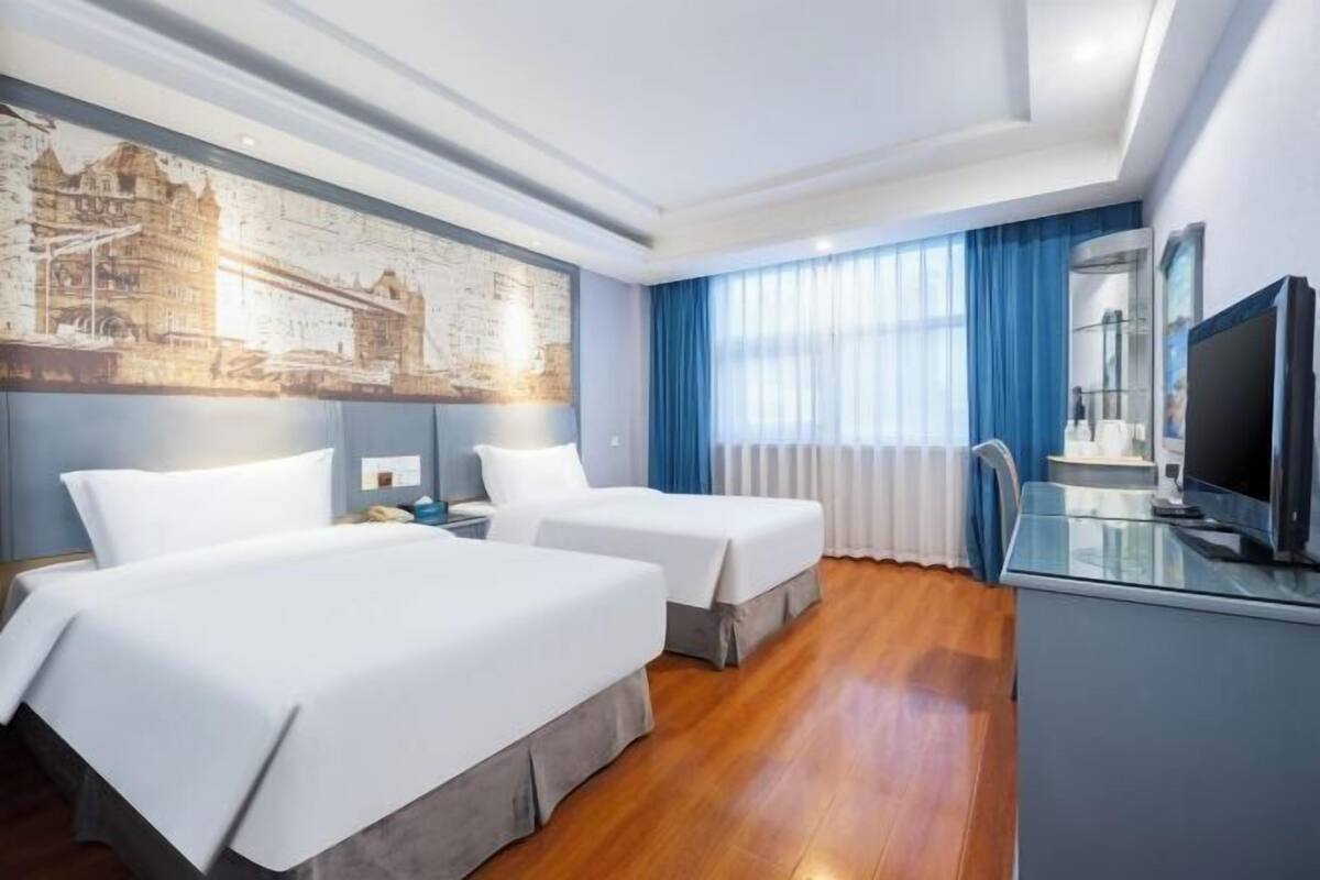Habitación de hotel con dos camas, televisor, escritorio y un ventanal grande con cortinas.