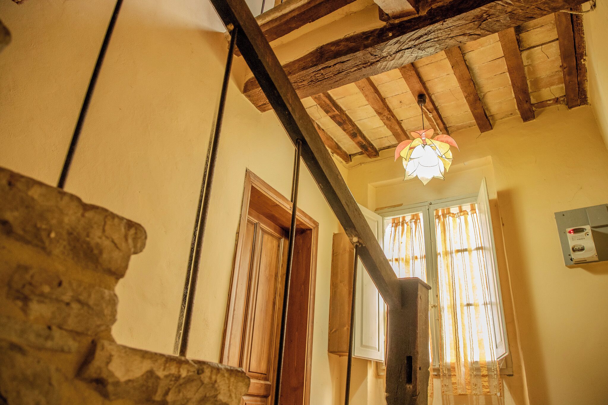Un escalier avec des poutres en bois et un mur en pierre, menant à une pièce dotée d’une fenêtre et de rideaux.