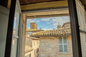 A view through a window of a narrow street with stone buildings, a bell tower, and a tiled roof. - Casa Vacanze Menta&Rosmarino (Mercatello sul Metauro)
