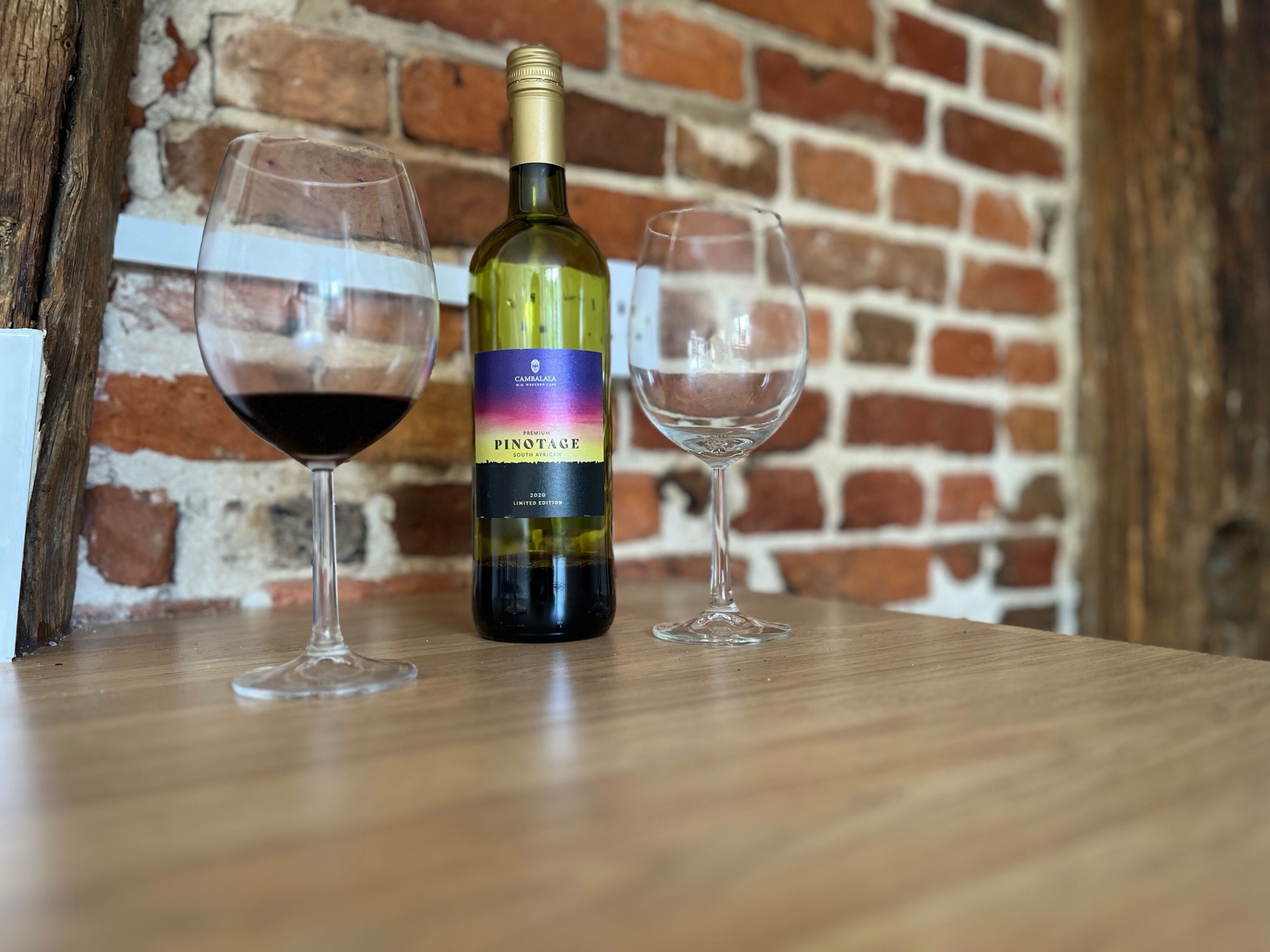 A bottle of wine and two glasses on a wooden table against a brick wall.
