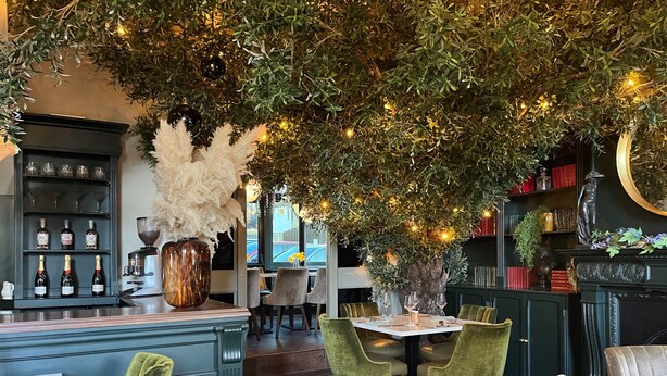 Een restaurant met een bar, groene beklede stoelen, een tafel met een vaas en een grote boom met lichtjes.