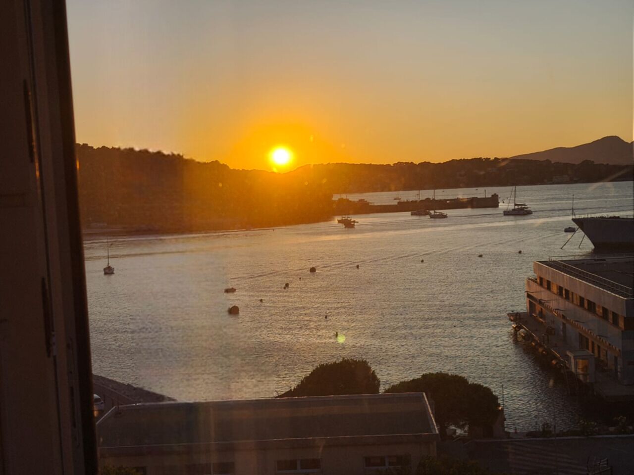 A sunset view from a window overlooking a body of water with boats and a distant shoreline.