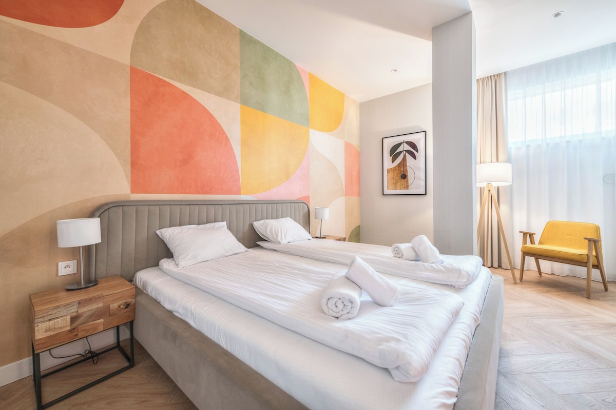 Habitación de hotel con dos camas, una mesita de noche de madera, una lámpara, una silla y un mural abstracto grande en la pared.