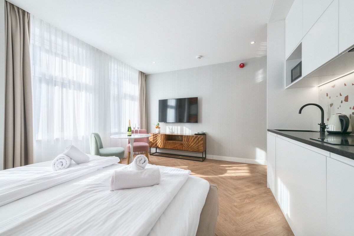 Una habitación de hotel moderna con una cama, una mesita con una botella y vasos, un televisor montado en la pared y una kitchenette.